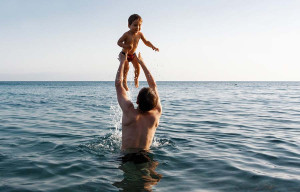 A man in the ocean throwing a happy baby up in the air. 