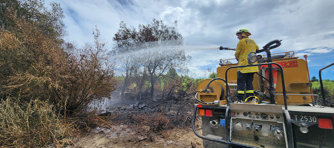Firefighter spraying water onto burnt vegetation from a fire truck