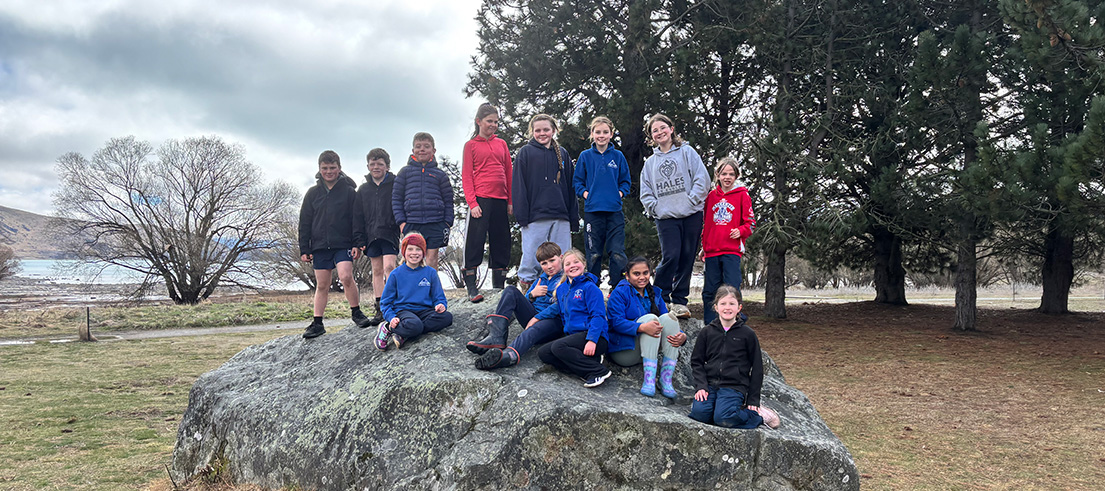 Group of school students gathered on a large rock beside trees and a lake in a rural landscape